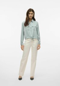 Camicia azzurro chiaro a maniche lunghe con due tasche sul petto e orlo con cordino, abbinata a pantaloni dritti crema e scarpe nere a punta.