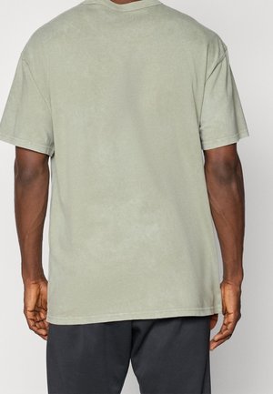 Mand iført lysegrøn kortærmet t-shirt set bagfra med mørke bukser mod en ensfarvet baggrund.