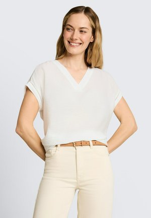 Lachende vrouw met schouderlang blond haar, gekleed in een wit T-shirt met V-hals en korte mouwen, ingestopt in beige broek met een lichtbruine riem.