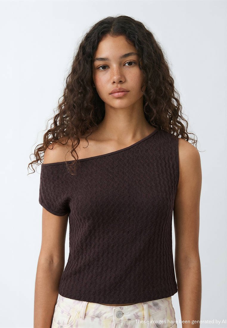 Jeune femme aux cheveux bouclés portant un haut asymétrique foncé et un pantalon clair à motifs, debout devant un fond uni clair.