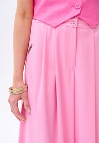 Un haut sans manches rose clair associé à un pantalon ample assorti. Présente un tissu lisse, des plis et un détail d'étiquette sur le pantalon.