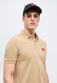 Νέος άνδρας που φοράει μπεζ πόλο πουκάμισο με κόκκινο λογότυπο στο στήθος και μαύρο καπέλο του μπέιζμπολ με κόκκινο σχέδιο, κοιτάζοντας μπροστά.