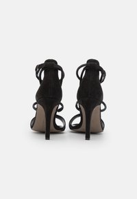 Sandalias de tacón alto de ante negro con parte trasera abierta, correas ajustables en el tobillo y recortes curvados en los lados. Suela interior suave.