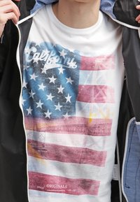 Personne portant un t-shirt blanc avec une impression fanée du drapeau américain et le mot « California », partiellement caché par une veste noire.