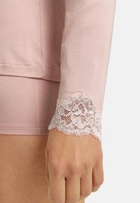 Primer plano de una mano y una muñeca que llevan una blusa de manga larga de color rosa claro con un borde de encaje floral, combinada con pantalones cortos rosa a juego.