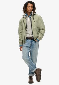 Lichtgroene bomberjack over een grijze hoodie en een blauwe shirt, gecombineerd met lichtblauwe jeans en bruine laarzen. Casual, gelaagde outfit met verschillende texturen.