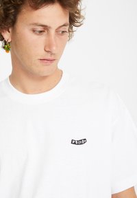 Weißes Baumwoll-T-Shirt mit rundem Halsausschnitt, versehen mit einem kleinen schwarzen gestickten Logo auf der linken Brust. Glatte Textur, kurze Ärmel.