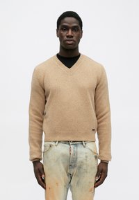 Suéter de cuello en V en beige, con tejido texturizado. Diseño ajustado con puños y dobladillo acanalados. Llevado con jeans claros desgastados. Branding mínimo visible.