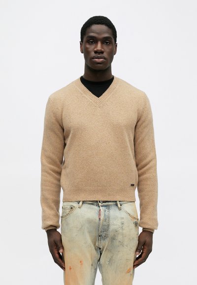 Beige Pullover mit V-Ausschnitt, strukturierter Strick. Enganliegendes Design mit gerippten Bündchen und Saum. Getragen mit hellblauen, ausgewaschenen Jeans. Minimale Markenkennzeichnung sichtbar.