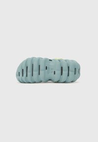 Helle blaue Schaumstoffsohle eines Sneakers mit einem wellenförmigen, strukturierten Muster und schwarzen Rillen. Verfügt über einen kleinen grünen Akzent in der Nähe der Ferse.