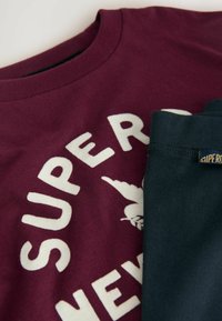 Felpa borgogna con lettering bianco, abbinata a pantaloni verde scuro con etichetta. Tessuto di cotone strutturato, design casual.