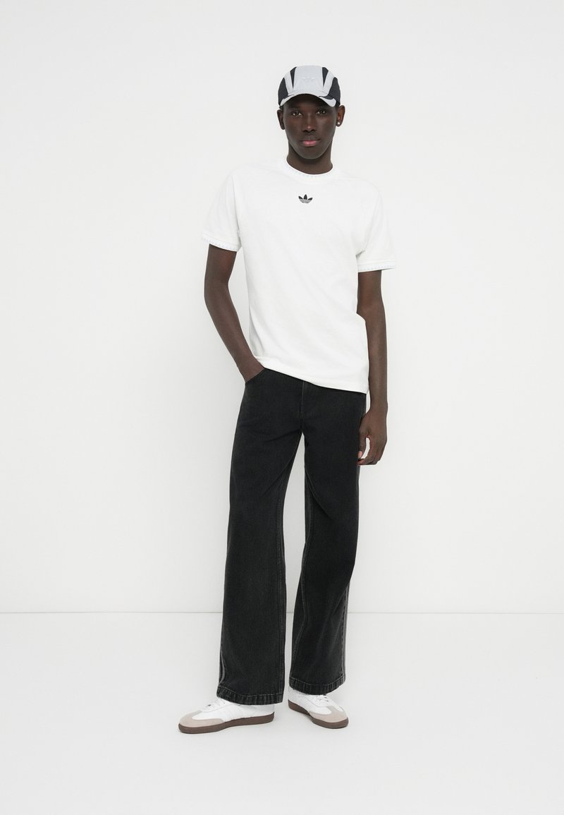 Maglietta bianca con piccolo logo nero, jeans neri a gamba larga e berretto grigio a righe. Le calzature hanno una base bianca con dettagli beige.