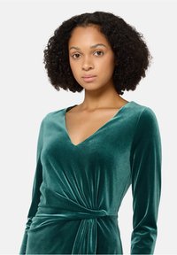 Emeraldgroene fluwelen jurk met een V-hals, lange mouwen en een gevlochten taille-detail, met een zachte glans en een gladde textuur.