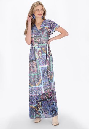 Kobieta w kolorowej sukience z wzorem, z krótkimi, falowanymi rękawkami i niebieskimi, wyrazistymi kolczykami, pozująca z dłonią przy twarzy.