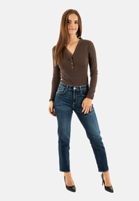 Top marron côtelé à manches longues avec accents de bouton, associé à un jean fitted bleu foncé. Modèle portant des escarpins noirs à bouts pointus, se tenant devant un fond blanc.