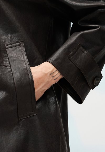 Καφέ δερμάτινο μπουφάν με μαλακή υφή, μανσέτα με κουμπιά και πλαϊνή τσέπη με το χέρι μέσα. Διακριτικές λεπτομέρειες ραφής εμφανείς.