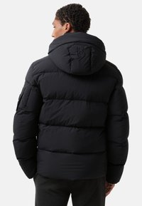 Man in een zwart gewatteerd pufferjack met capuchon, voorzien van een gestikt "B"-logo, met de rug naar de camera tegen een effen achtergrond.
