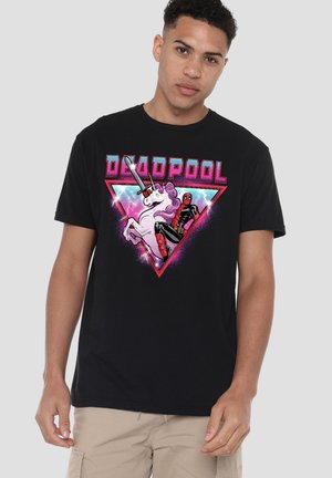 Young man wearing black t-shirt with colorful Deadpool riding a white unicorn graphic and pink triangle background.