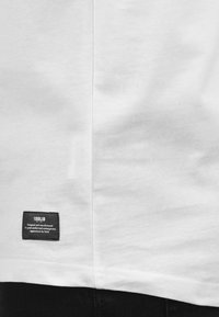 Weißes Baumwoll-T-Shirt mit einer Seitennaht und einem schwarzen Etikett mit weißem Text, das sich in der Nähe des Saums befindet. Glatte Textur und schlichtes Design.