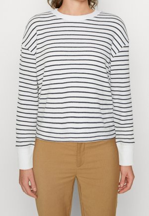 Personne portant un haut blanc à manches longues avec des rayures noires horizontales et un pantalon beige, debout devant un fond uni clair.