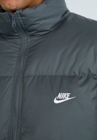 Hallide hallhübedejoond puffer-jakk tumedat, lukuga ja vasakpoolse rinna valge Nike'i logoga. Sile, kerge kangas, millel on tikitud osad.