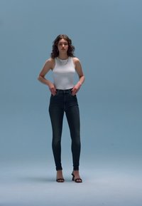 Λευκό αμάνικο τοπ, σκούρο στενό τζιν και μαύρα ψηλοτάκουνα. Το μοντέλο στέκεται μπροστά σε φωτεινό γαλάζιο φόντο, παρουσιάζοντας μια εφαρμοστή σιλουέτα.