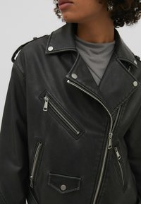 Veste de motard en cuir noir avec quincaillerie argentée, plusieurs poches zippées et détails à pression. Dotée d'un large col et d'un léger éclat.