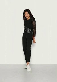 Conjunto preto com um top justo com mangas longas em renda e calças de perna reta. Inclui uma mala de cintura preta com detalhe em corrente.