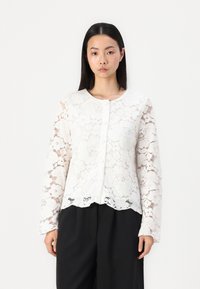 Blusa bianca in pizzo con motivi floreali, maniche lunghe e scollo rotondo con chiusura anteriore a bottoni. Dettaglio con orlo ondulato.