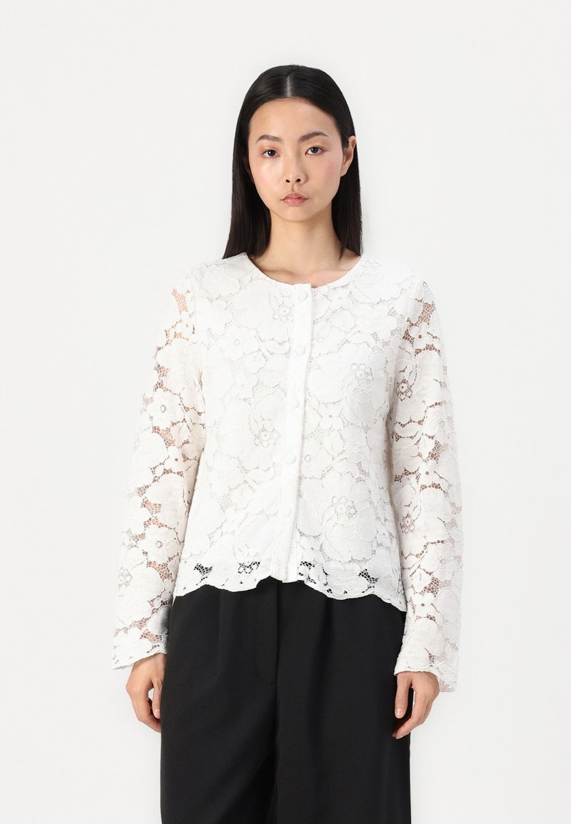 Blusa bianca in pizzo con motivi floreali, maniche lunghe e scollo rotondo con chiusura anteriore a bottoni. Dettaglio con orlo ondulato.