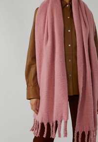 Écharpe rose pâle à franges longues, douce, drapée sur une chemise en cuir marron à boutons et un pantalon foncé, portée par une personne sur un fond uni.