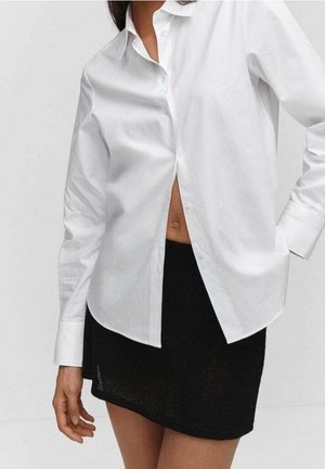 Kvinde iført en hvid skjorte med knapper, delvist åben ved taljen, og en sort mininederdel, stående mod en ensfarvet baggrund.