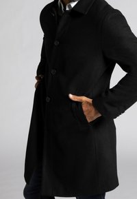 Cappotto in misto lana nero con colletto, bottoni frontali e tasche laterali. Vestibilità da sartoria e texture liscia. Dettaglio di cucitura visibile sulle maniche.