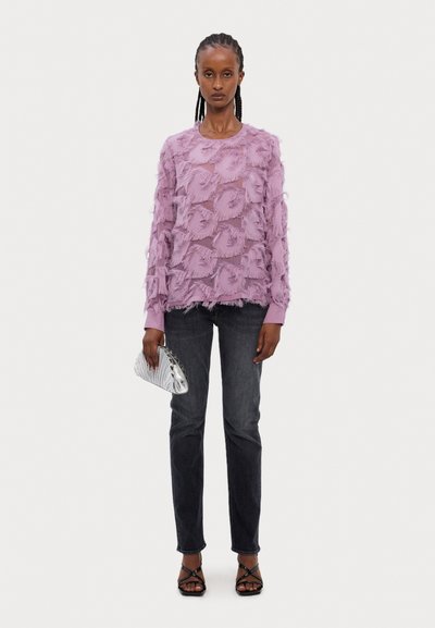 Top de manga larga lila con patrón texturizado y volantes; combinado con jeans oscuros y un bolso metálico; sandalias negras de tiras completan el look.