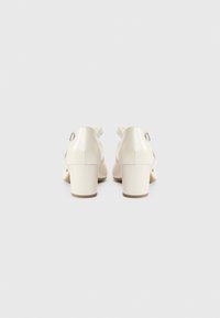 Κρεμ ψηλοτάκουνα παπούτσια με λείο δερμάτινο φινίρισμα, λουρί στον αστράγαλο και τετράγωνο τακούνι. Η οπίσθια όψη αναδεικνύει το λουρί και τον δομικό σχεδιασμό.