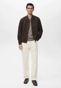 Braune Wildleder-Bomberjacke über einem beigen Hemd, kombiniert mit weißen Hosen und braunen Schuhen. Glatte Textur, minimalistisches Design, neutrale Farben.