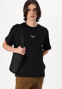 Schwarzes Baumwoll-T-Shirt mit weißem Textlogo in der Mitte. Kombiniert mit einer kleinen schwarzen Stoff-Umhängetasche. Model trägt hellbraune Cordhosen.