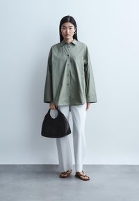 Žena stojící a nosící zelenou volnou košili s knoflíky, bílé kalhoty s výšivkou na lemech, hnědé sandály a držící černou kabelku.