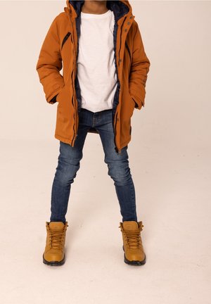 Enfant portant un manteau d'hiver orange à capuche, une chemise blanche, un jean bleu et des bottes lacées beige, debout les mains dans les poches du manteau.