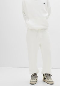 White hoodie with a front pocket, paired with matching loose-fit pants. Notable for its soft texture and simple design. Worn with gray sneakers.