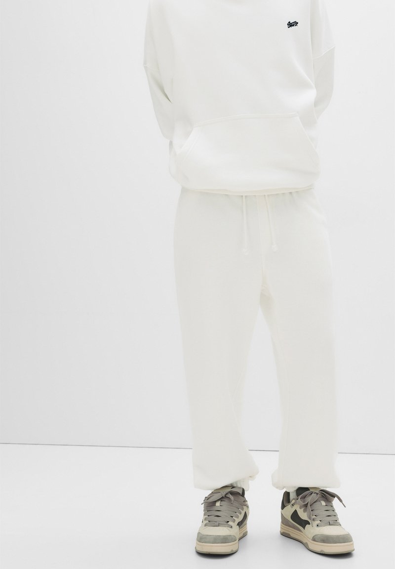 White hoodie with a front pocket, paired with matching loose-fit pants. Notable for its soft texture and simple design. Worn with gray sneakers.