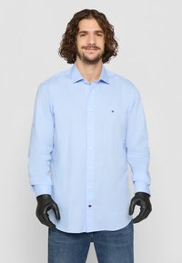 Lys blå skjorte med knapper og lange ermer, laget av glatt stoff. Har en liten logo detalj på brystet, kombinert med mørke jeans.