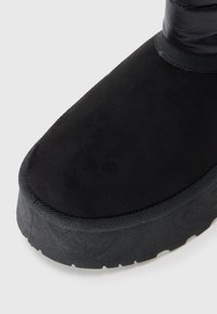 Botte en daim noir avec une semelle en caoutchouc texturée et plate. Présente un empiècement rembourré avec des accents élastiques près de la cheville. Fini lisse.