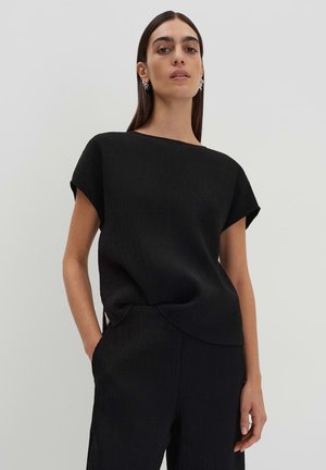 Black short-sleeve top with a boat neckline, textured fabric, and an asymmetrical hem. Worn with matching black trousers.