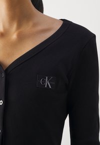 Musta nyöritetty kangas toppi V-aukolla, jossa on napp closure ja suorakulmainen Calvin Klein Jeans logo -tarran valmistuksessa rintaosassa.