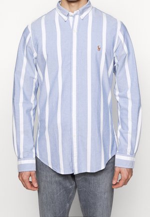 Homme portant une chemise bleu clair à manches longues avec de larges rayures blanches verticales et un jean gris, debout devant un fond uni.