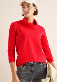 Rotes Langarmoberteil mit schicker Kapuze, aus glattem Stoff gefertigt. Kombiniert mit grauen Jeans, die einen lässigen Schnitt und lockere Passform zeigen.