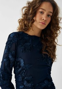 Bluză lungă, cu mânecă lungă, de culoare bleumarin, cu broderie florală texturată, având un decolteu rotund și un material moale, transparent. Părul creț este vizibil în prim-plan.