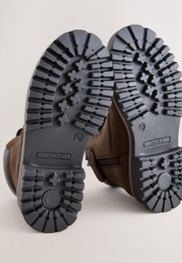 Braune Wildlederstiefel mit einer schwarzen Gummilaufsohle, die ein tiefes Profilmuster aufweist. Die Fersen- und Zehenbereiche haben abgerundete Kanten für besseren Halt.