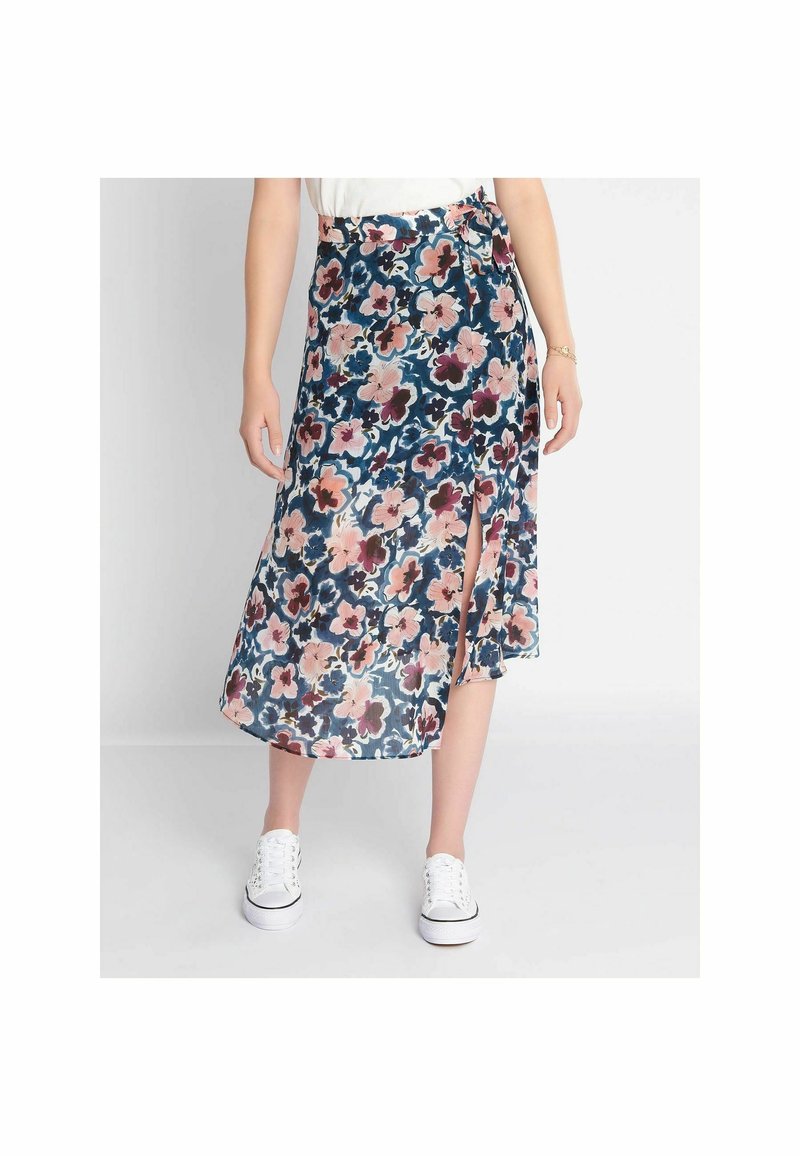 Femme portant une jupe portefeuille midi fleurie avec une fente latérale, associée à des baskets blanches et un haut blanc, debout devant un fond uni.